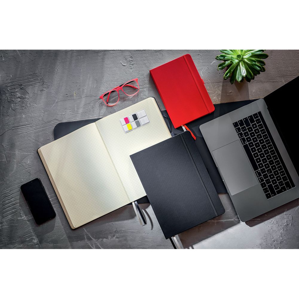 Ein offenes Notizbuch, ein rotes und ein schwarzes Notizbuch, Brille, Handy, Laptop und Pflanze.