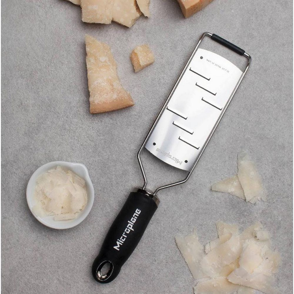 Käsemesser mit schwarzen Griff, daneben Käsebrocken und geriebener Käse.
