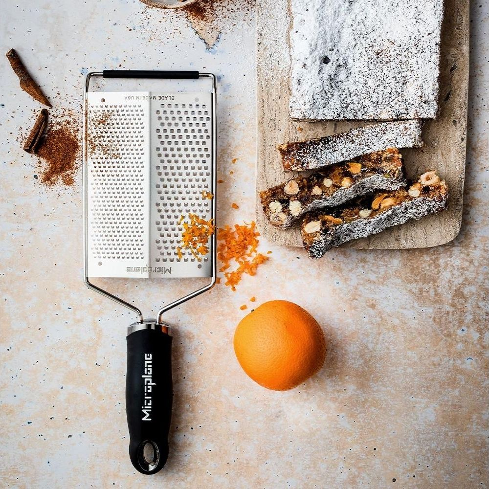 Ein Grater mit Orangenschale, Zimtstangen und Kuchenstücken auf einem Brett.