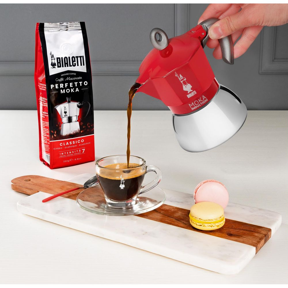 Czerwony moka pot Bialetti nalewa kawę do filiżanki. Obok paczka kawy.