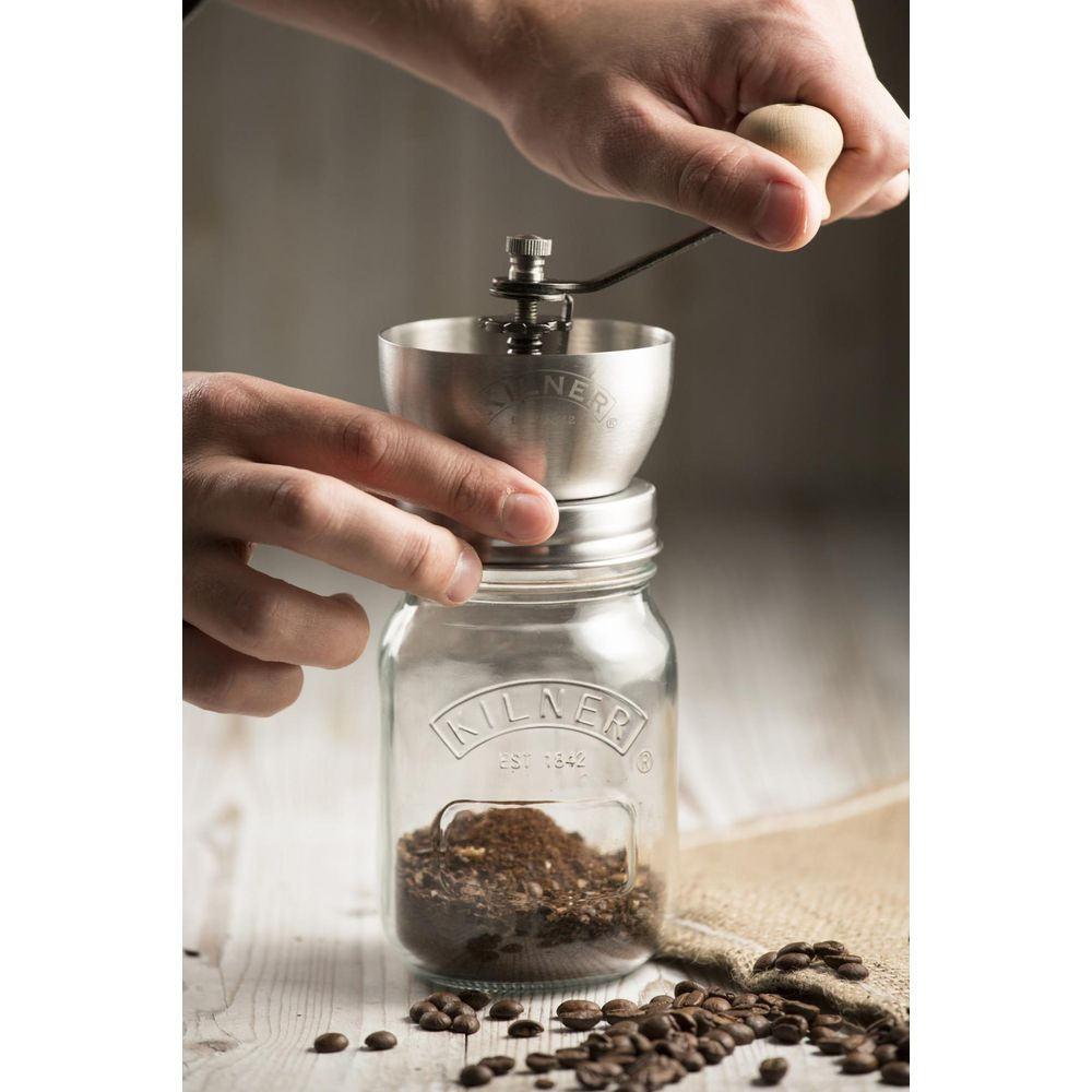 Person mahlt Kaffee mit einer Metallkaffeemühle in ein Glas. Kaffeebohnen liegen daneben.