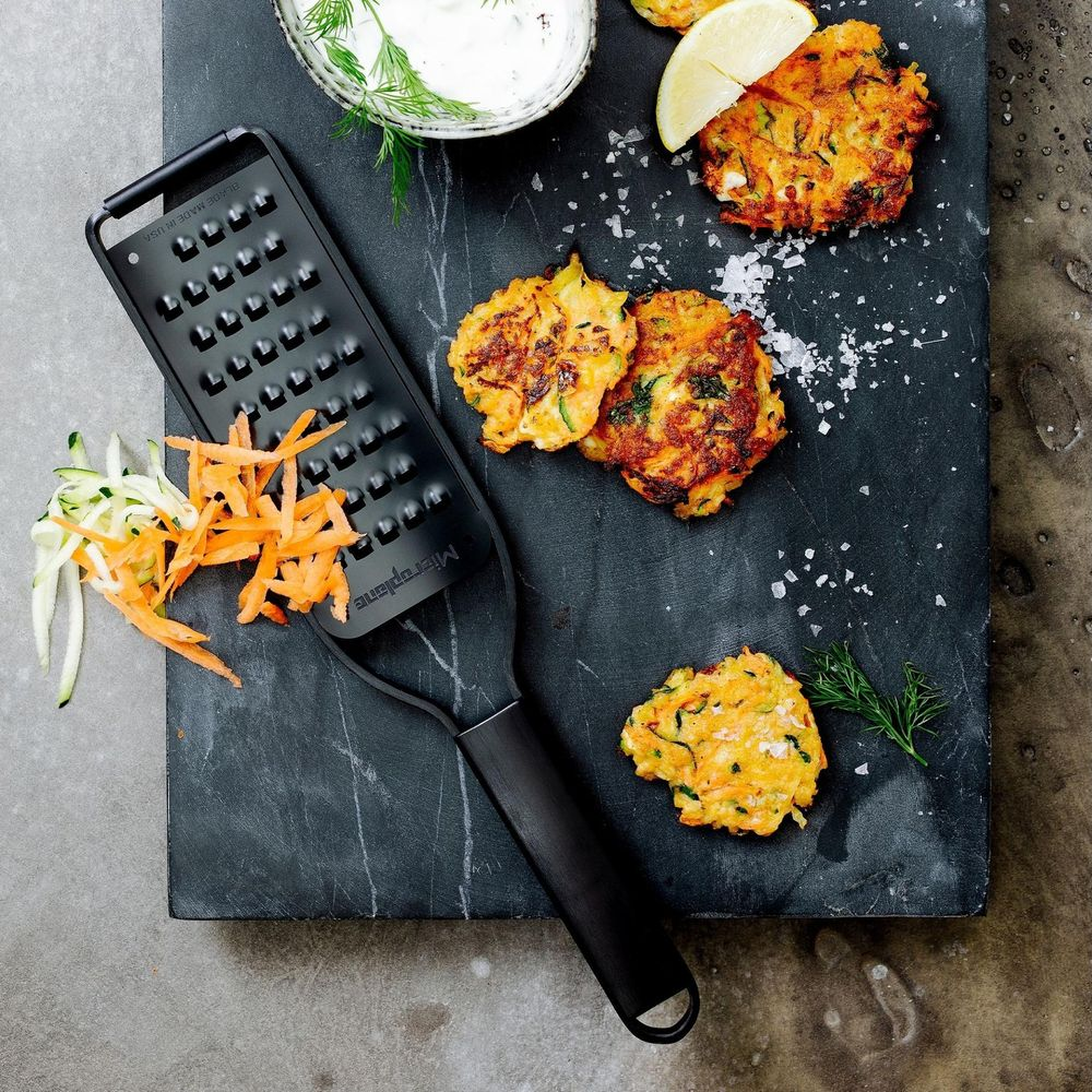 Puffer mit Reibe, geriebenen Karotten, Zucchini, Dill und Dip auf dunklem Schiefer.