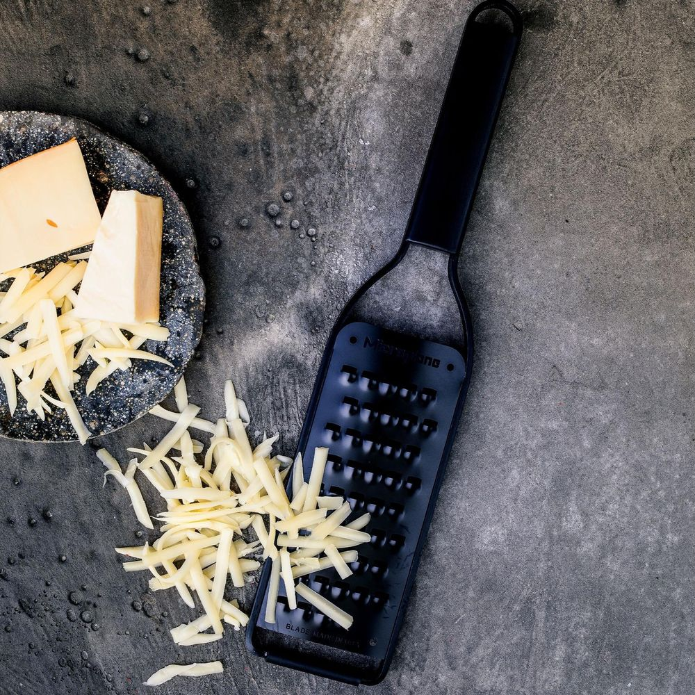 Ein schwarzer Käsereibe mit geriebenem und blockigem Käse auf grauer Oberfläche.
