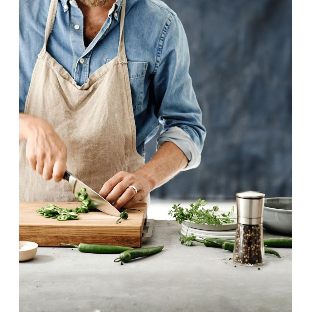 Mężczyzna w fartuchu kroi zieloną paprykę na desce, z młynkiem i ziołami.