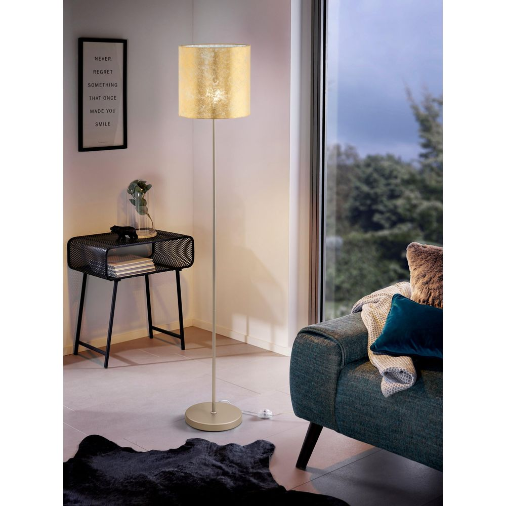 Un lampadaire à abat-jour doré est à côté d'un canapé. Une table d'appoint et un tableau à gauche.