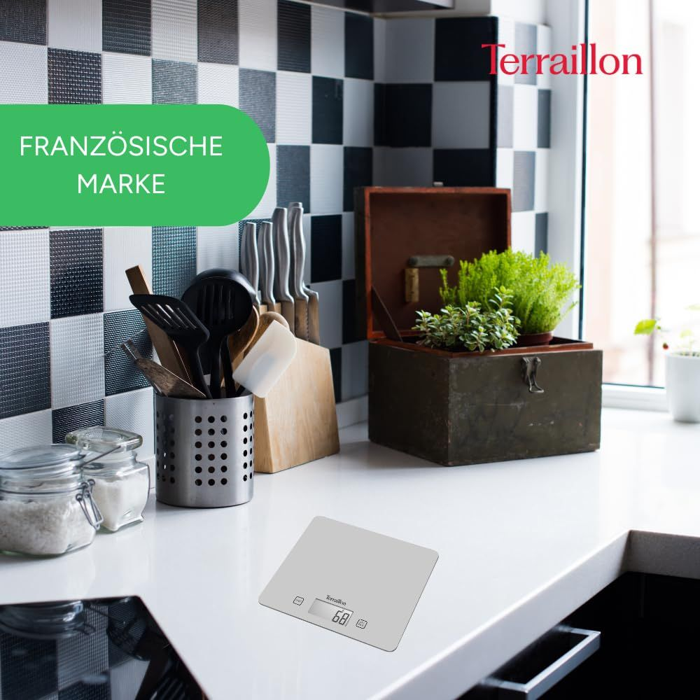 Bilancia da cucina grigia su un piano di lavoro con utensili e piante.