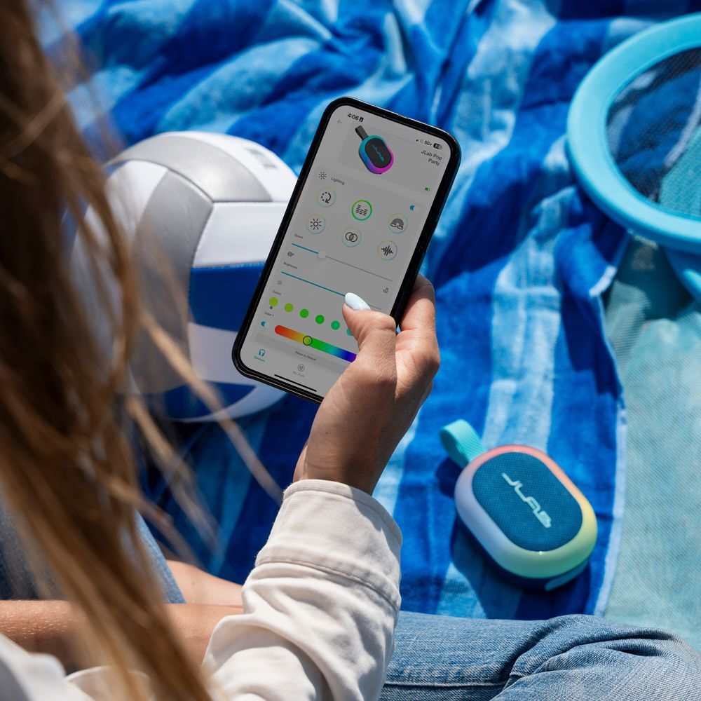 Personne utilise application mobile pour contrôler enceinte colorée. Ballon volley et serviette à proximité.