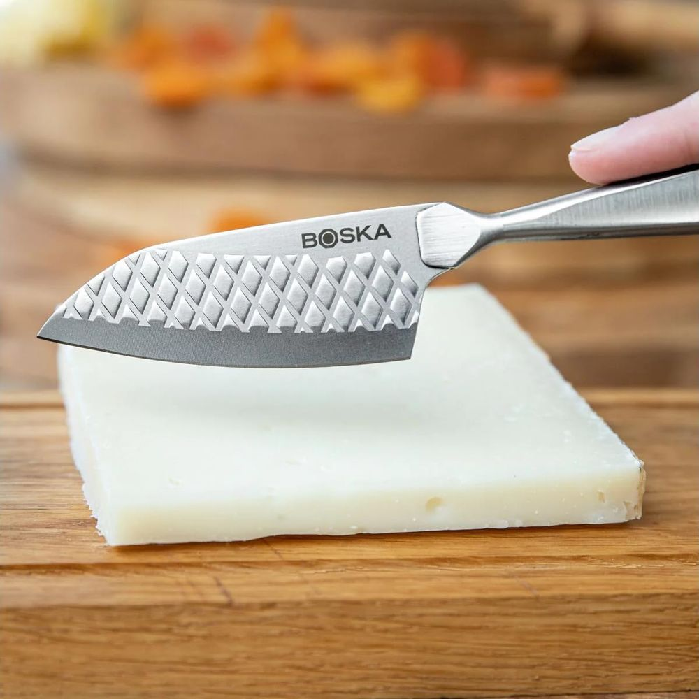 A Boska cheese knife with a diamond pattern cuts a slice of white cheese on a wooden board.