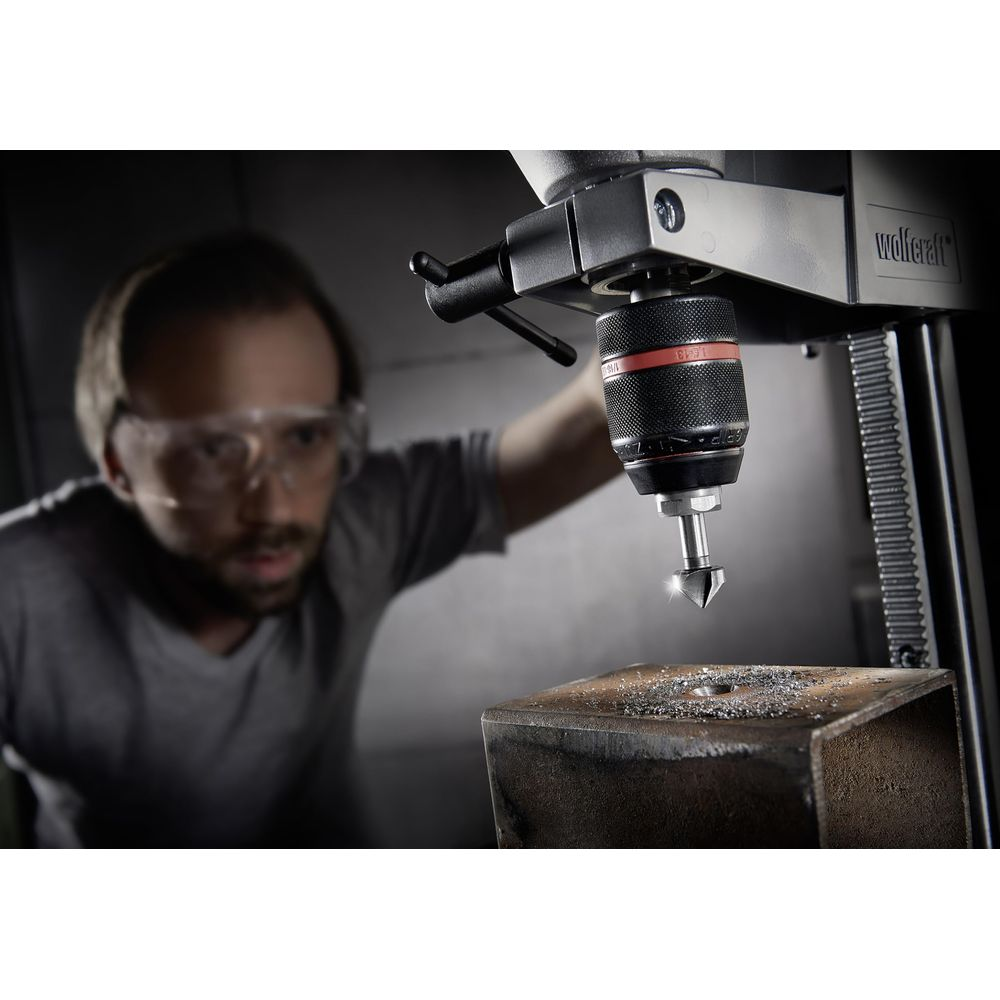 Hombre con gafas de seguridad taladra metal con taladro de columna, vuelan virutas.