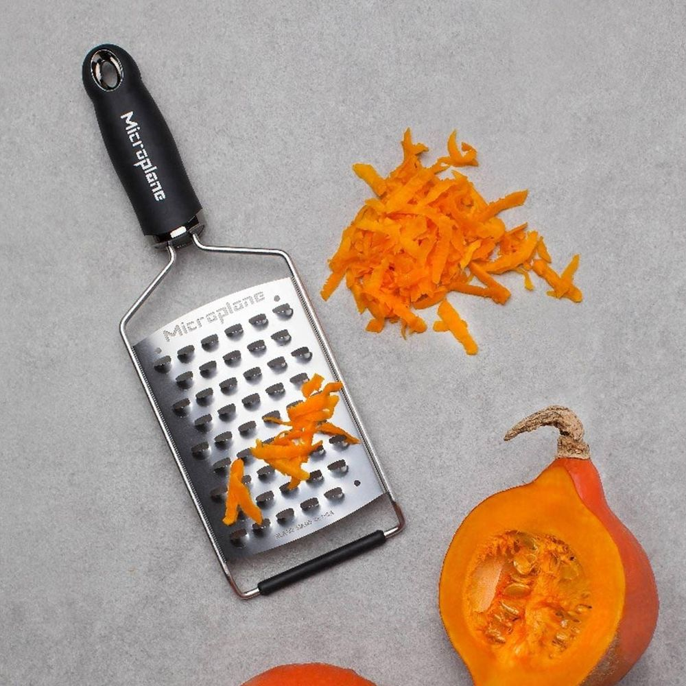 Ein Grater mit Orangenspalten und geriebenem Kürbis auf grauem Untergrund.