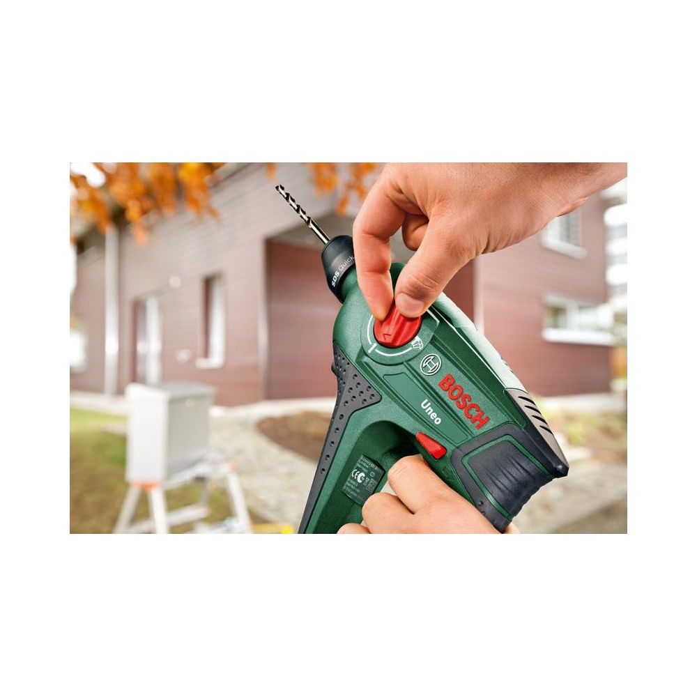 Ręka trzyma zielony wiertarkę Bosch z czerwonym przyciskiem przed domem.