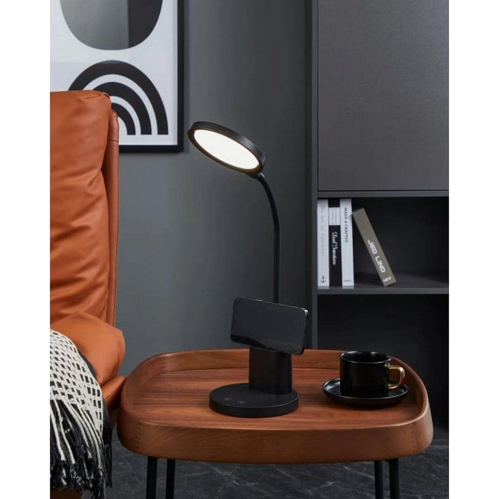 Eine moderne schwarze Schreibtischlampe mit Handyhalter steht auf einem Holztisch neben einer Kaffeetasse.
