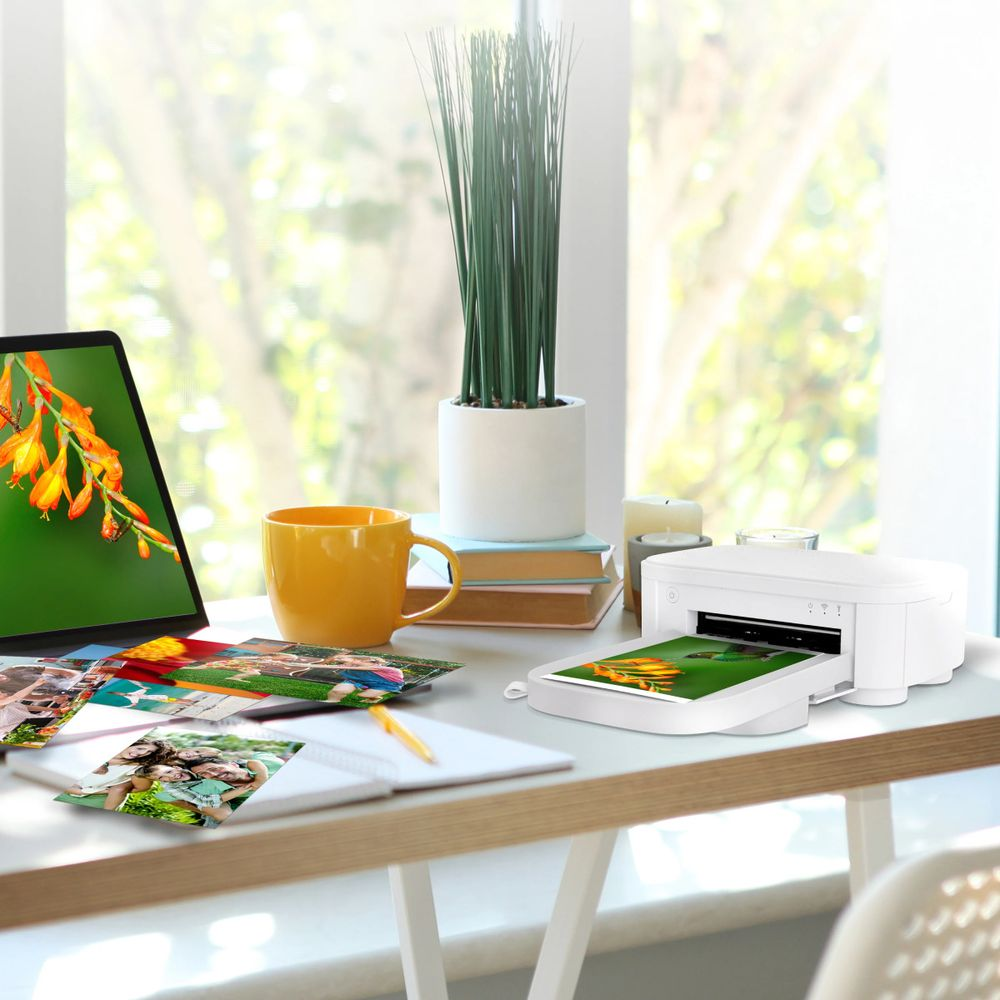 Biała drukarka drukuje obraz na biurku obok laptopa, kubka i zdjęć.