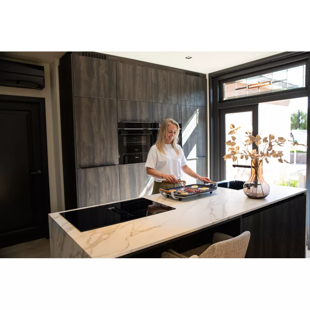 A woman fries eggs and bacon on a tabletop grill in a modern kitchen.