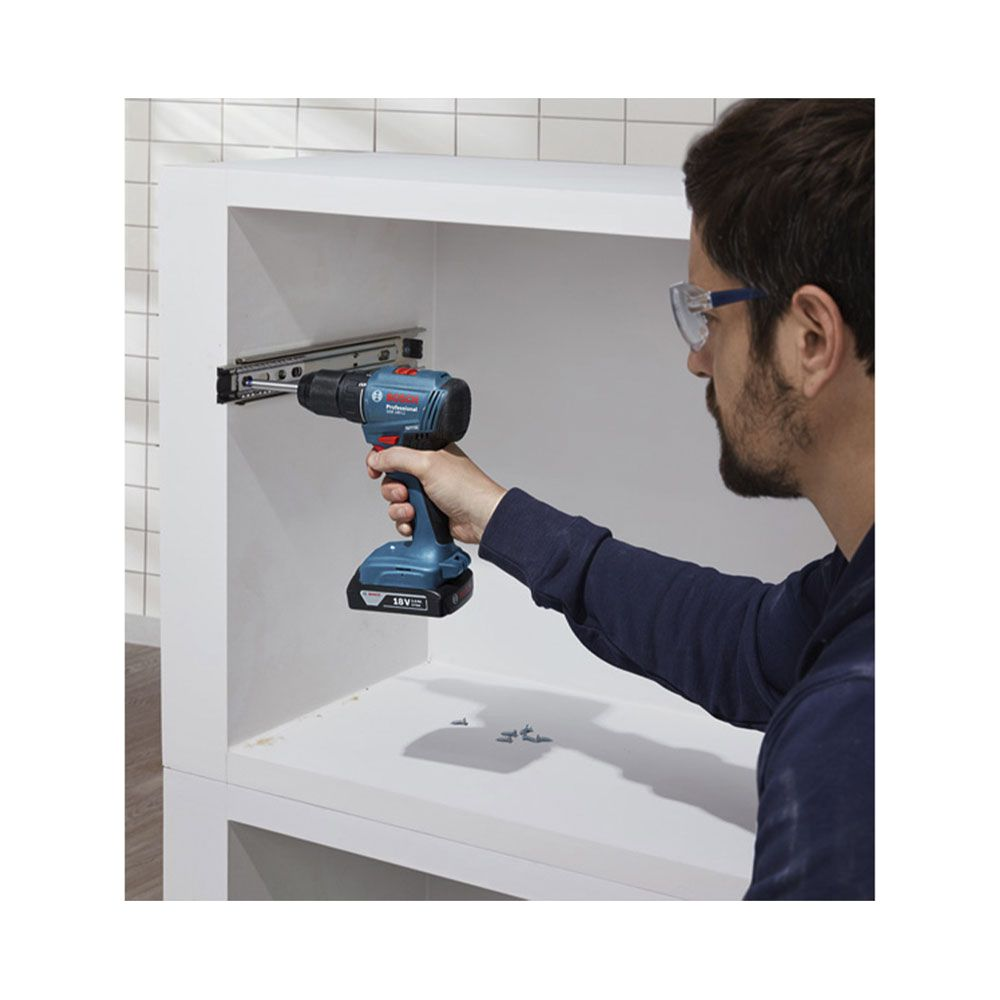 Mężczyzna wierci w białej powierzchni niebieską wiertarką. Okulary.