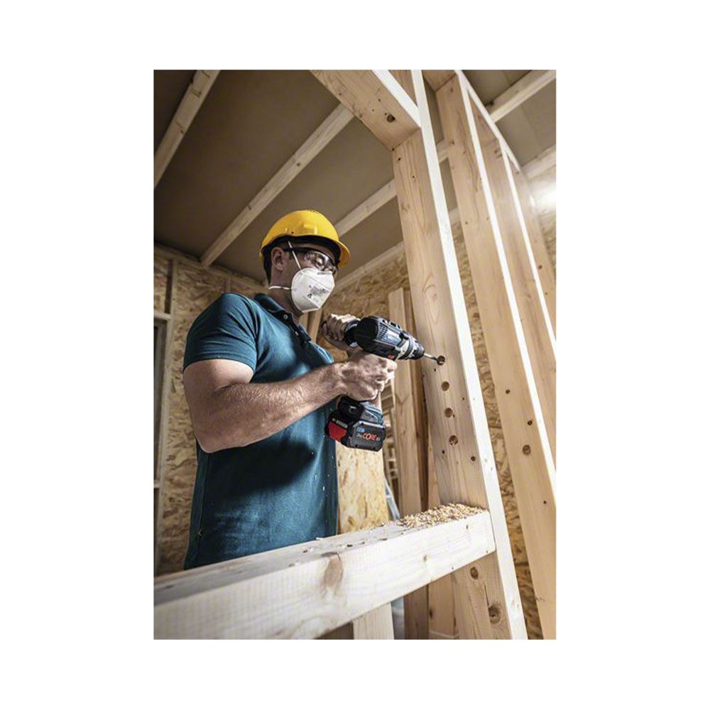 Mężczyzna w kasku wierci w drewnianej ramie, nosi maskę i okulary.