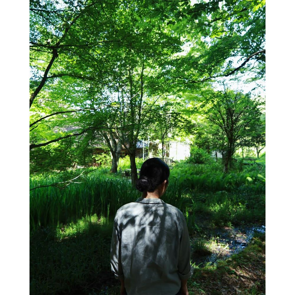 Une personne aux cheveux sombres en chignon regarde loin, portant une chemise grise dans une forêt verte luxuriante.