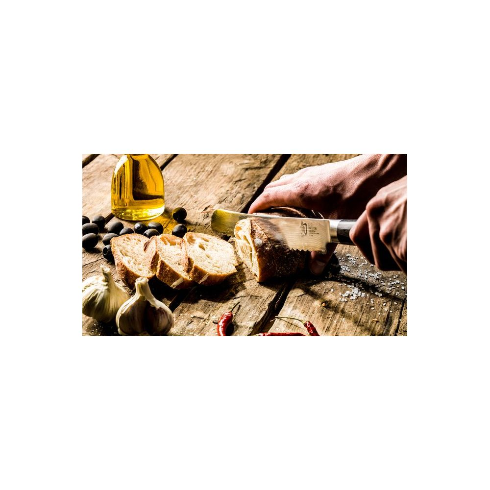 Ręce krojące chleb nożem, obok oliwek, czosnku, chili, oleju.