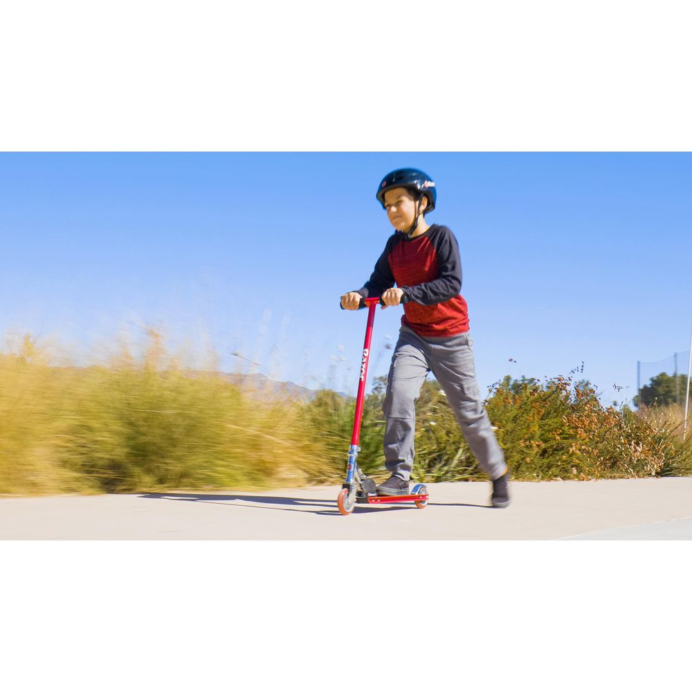 Ein Junge mit Helm fährt einen roten Roller auf einem Gehweg.