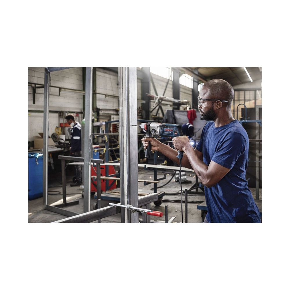 Mężczyzna wierci w metalowej ramie w warsztacie. W tle pracuje inny mężczyzna.