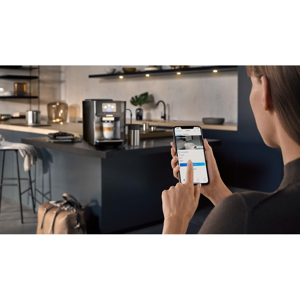 Kobieta steruje ekspresem do kawy smartfonem w nowoczesnej kuchni.