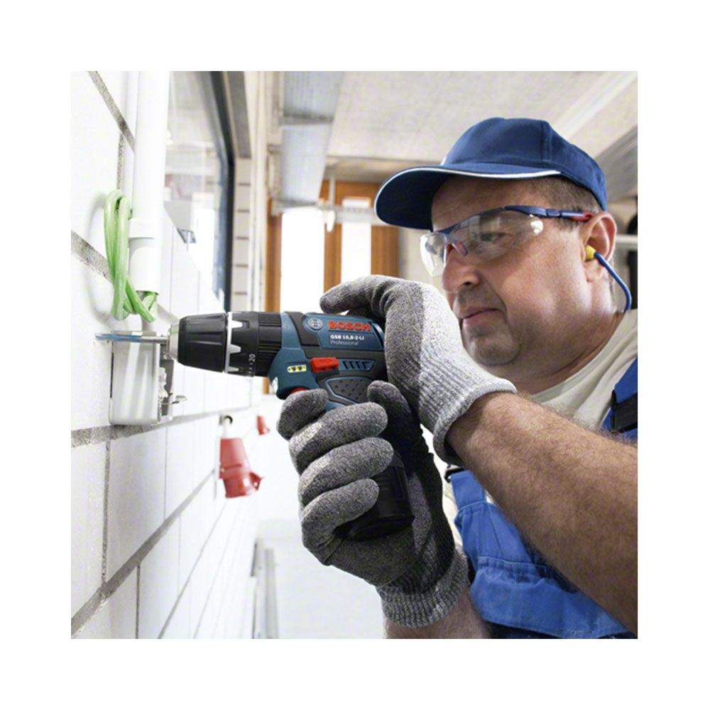 Mann mit blauer Kappe und Schutzbrille benutzt Akkuschrauber an weißer Wand.