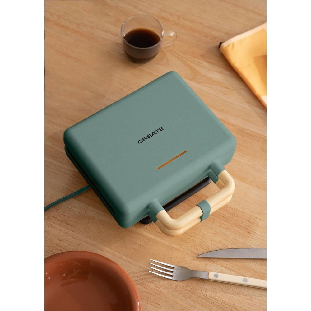 Prensa de sándwiches verde con mango de madera, taza de café, plato y cubiertos.