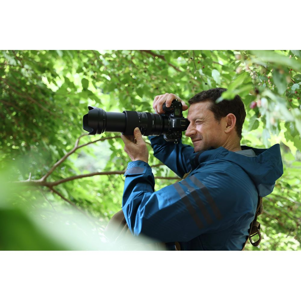 Mężczyzna z aparatem w lesie, w niebieskiej kurtce, otoczony zielonymi liśćmi.