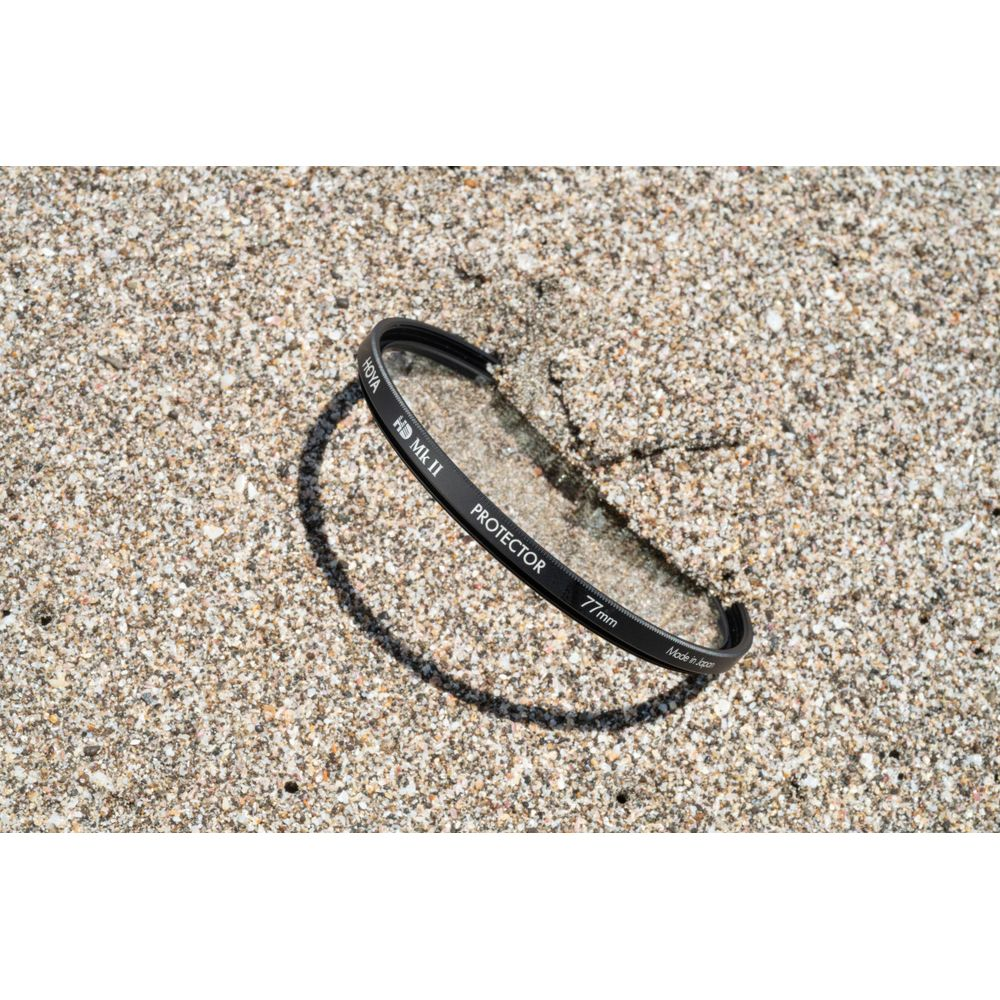 Ein Hoya HD Mk II 77mm Schutzfilter liegt auf Sand. Schatten fällt darauf.