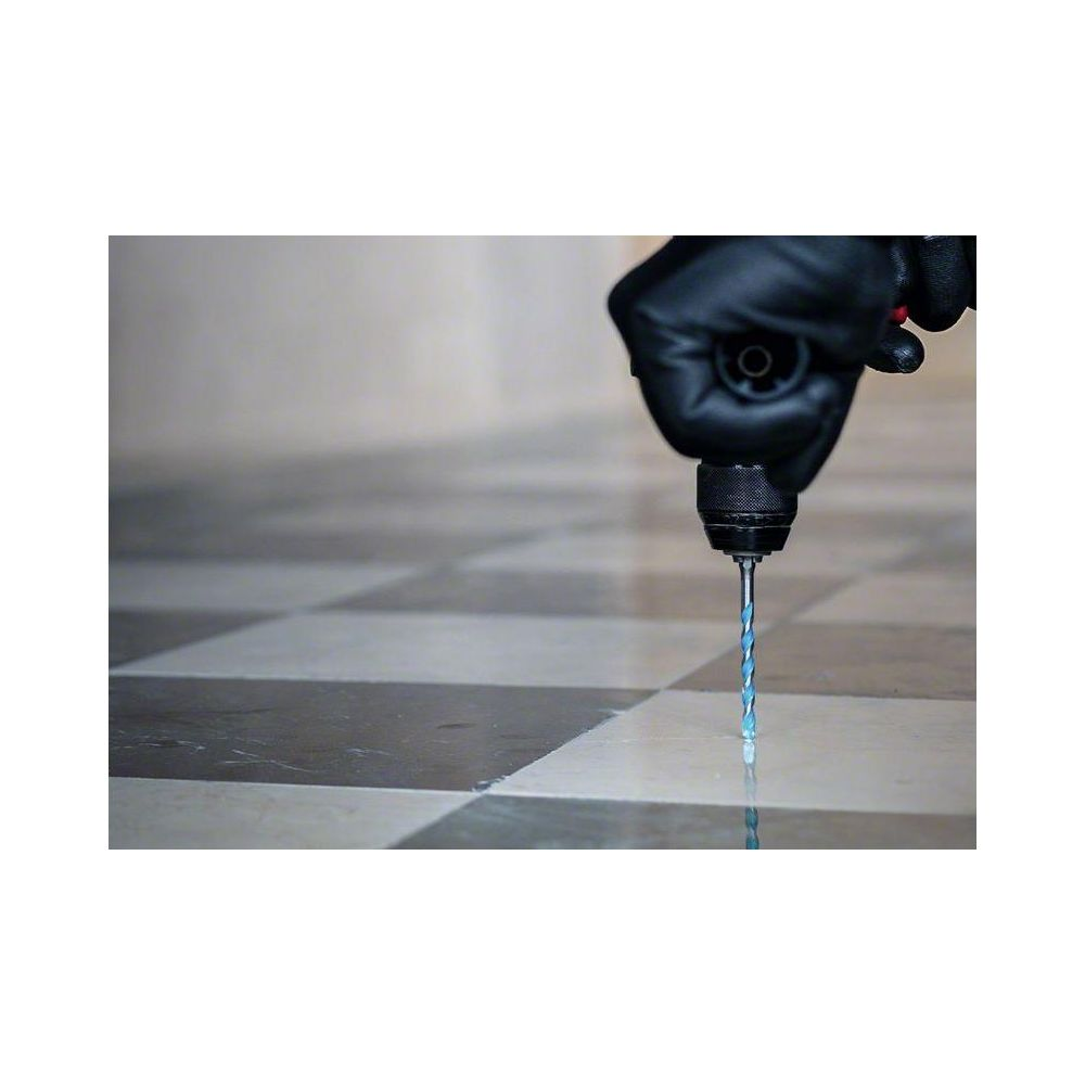 Ręka w czarnej rękawicy trzyma wiertarkę nad podłogą w kratkę.