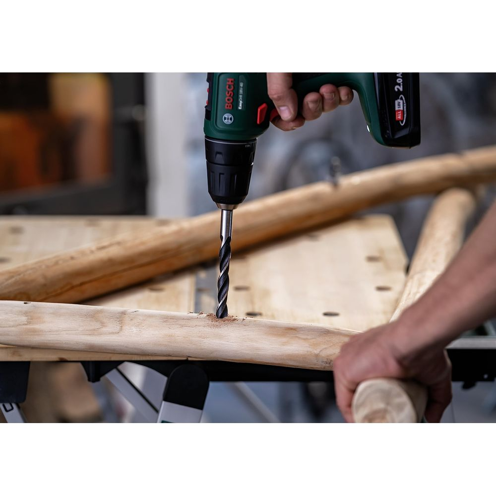 Perceuse Bosch verte troue poutre en bois, mains tenant la pièce.