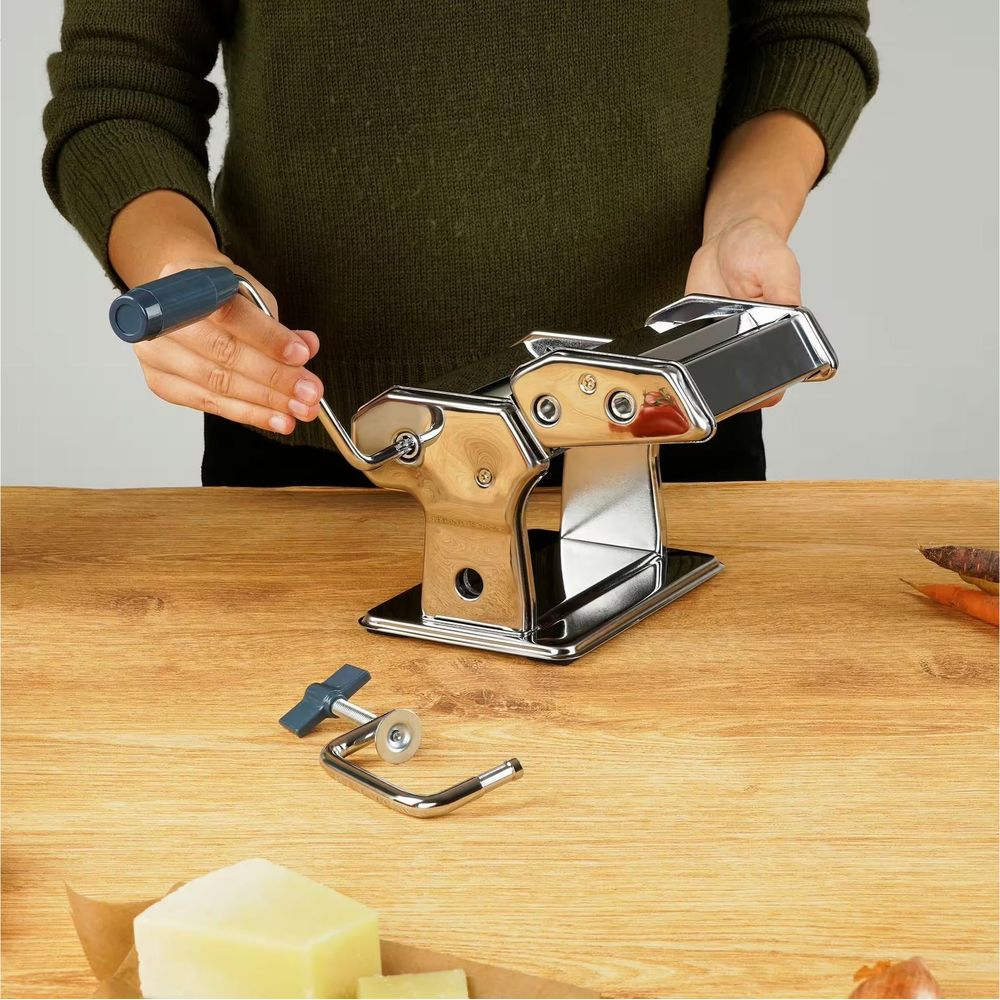 Person benutzt eine glänzende Nudelmaschine mit Kurbel auf einem Holztisch.