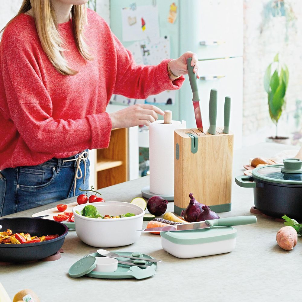 A woman in a red sweater places a knife into a wooden block. Kitchen utensils are on the counter.