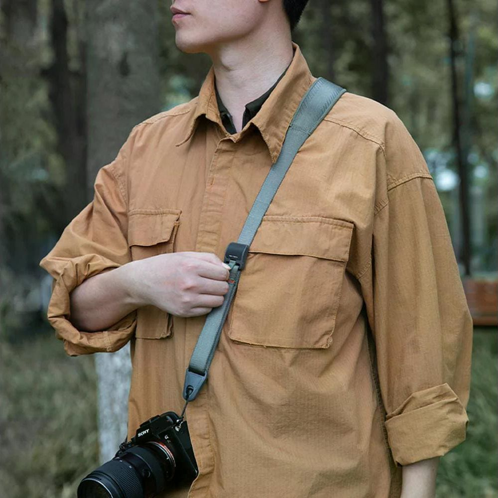 Mężczyzna w brązowej koszuli z paskiem i aparatem, na zewnątrz.