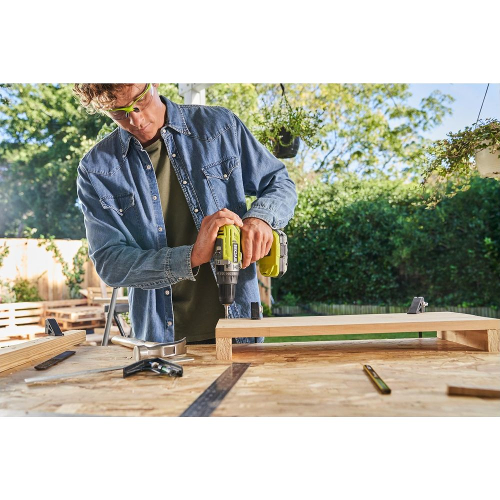 Mann in Jeanshemd und Schutzbrille bohrt Holz auf einem Tisch im Freien.