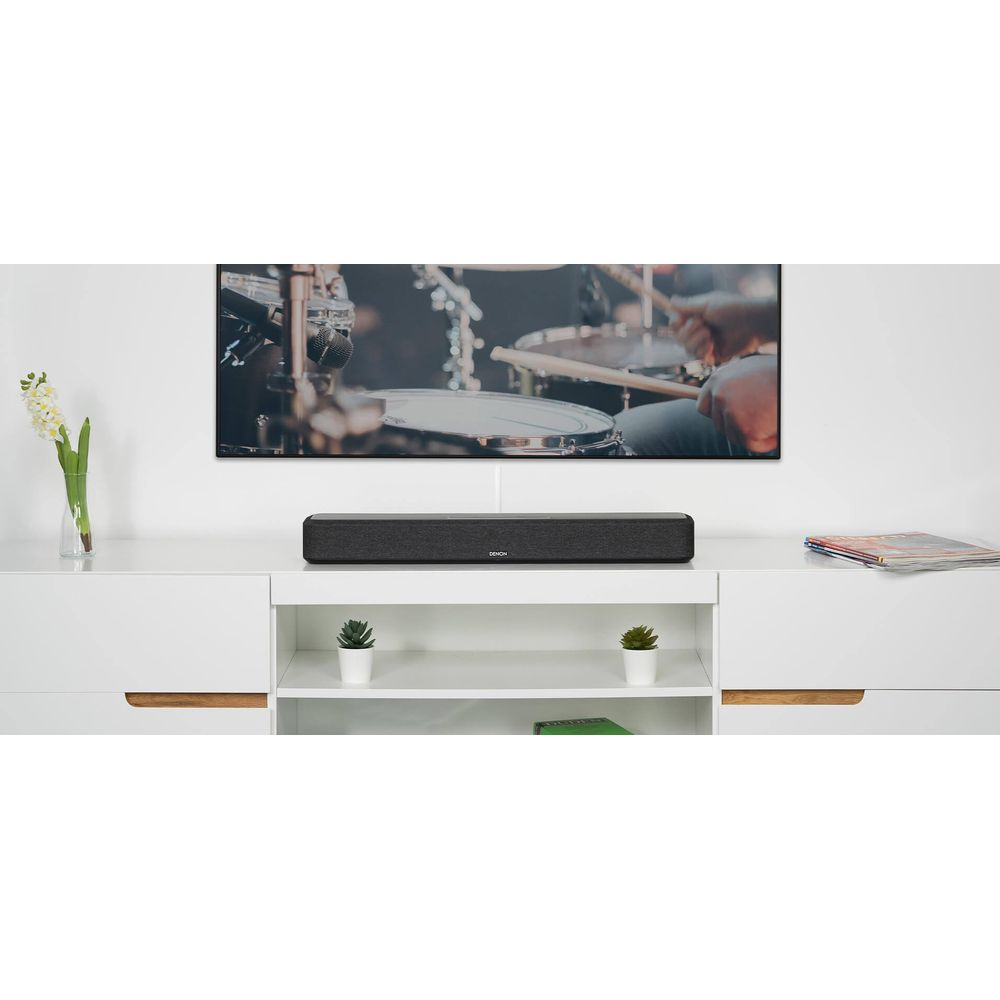 A dark soundbar sits on a white shelf in front of a TV showing a drummer.