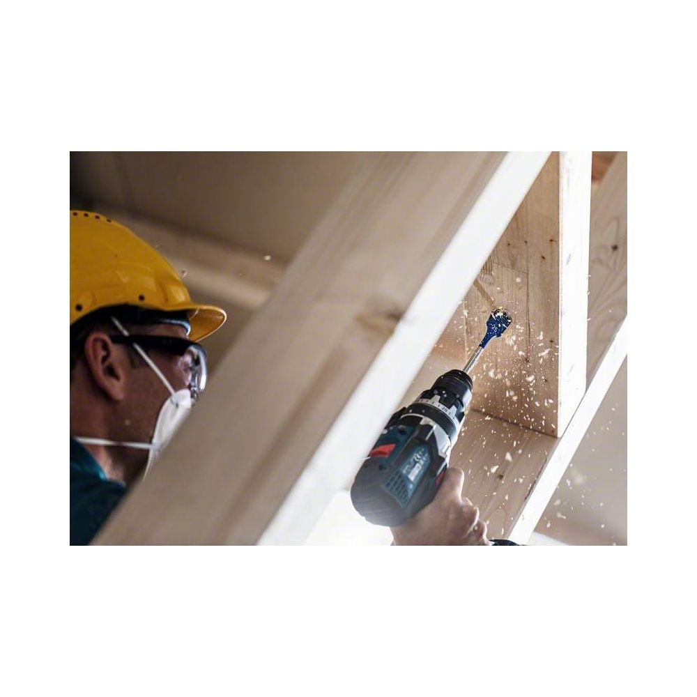 Mężczyzna w kasku wierci w drewnie, lecą wióry.