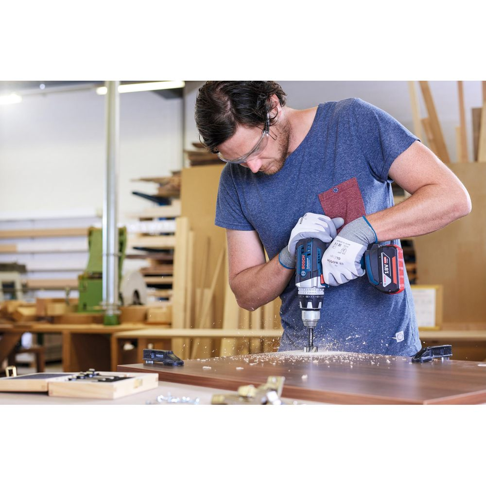 Mężczyzna wierci w drewnie wiertarką akumulatorową w warsztacie. Okulary, rękawice.