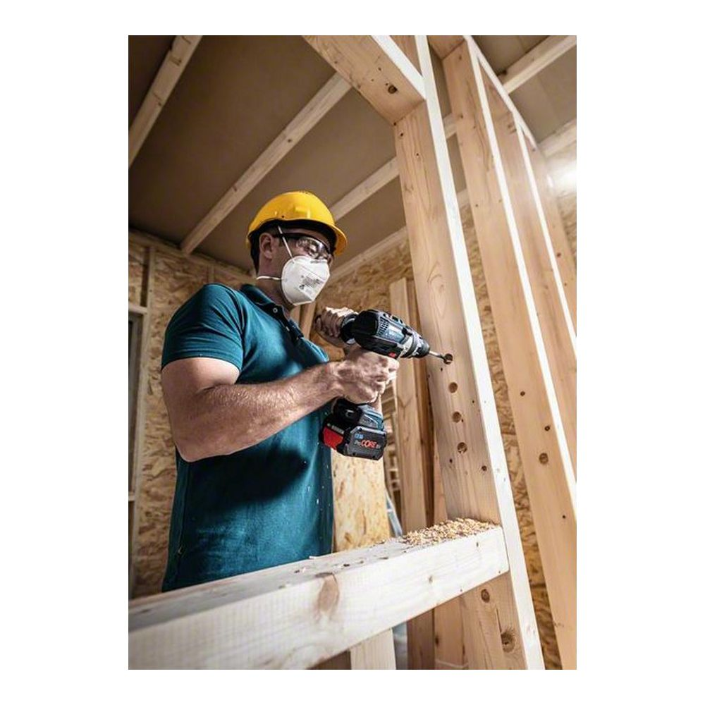 Mężczyzna w kasku i masce wierci w drewnianej ramie. Pył drzewny spada.