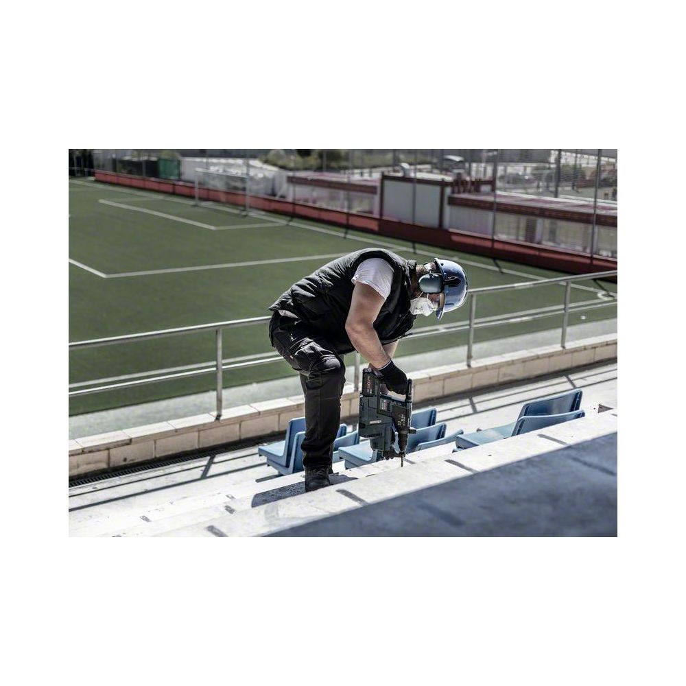 Mężczyzna z wiertarką na trybunach, boisko sportowe w tle.