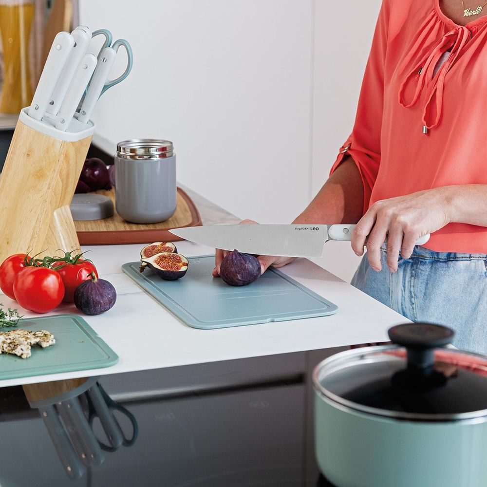 A person cuts a fig on a blue board with tomatoes and knives nearby.