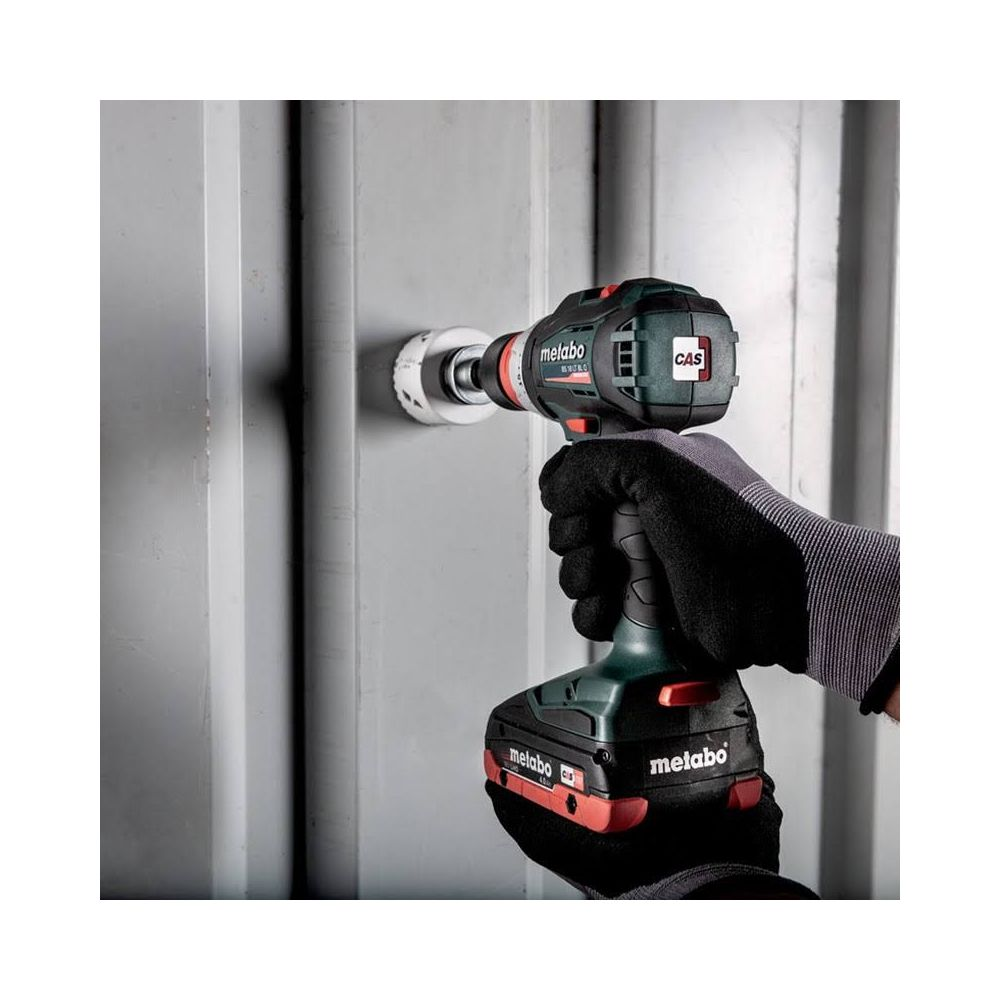 A gloved hand uses a cordless drill with a hole saw to drill into a grey corrugated wall.