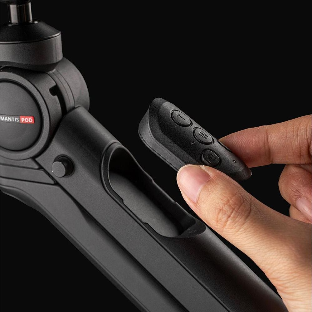 Une main tient une petite télécommande avec des boutons, pointant vers un trépied noir.