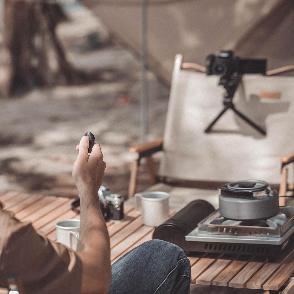Homme tient télécommande; réchaud camping, appareil photo, tasses sur table bois.