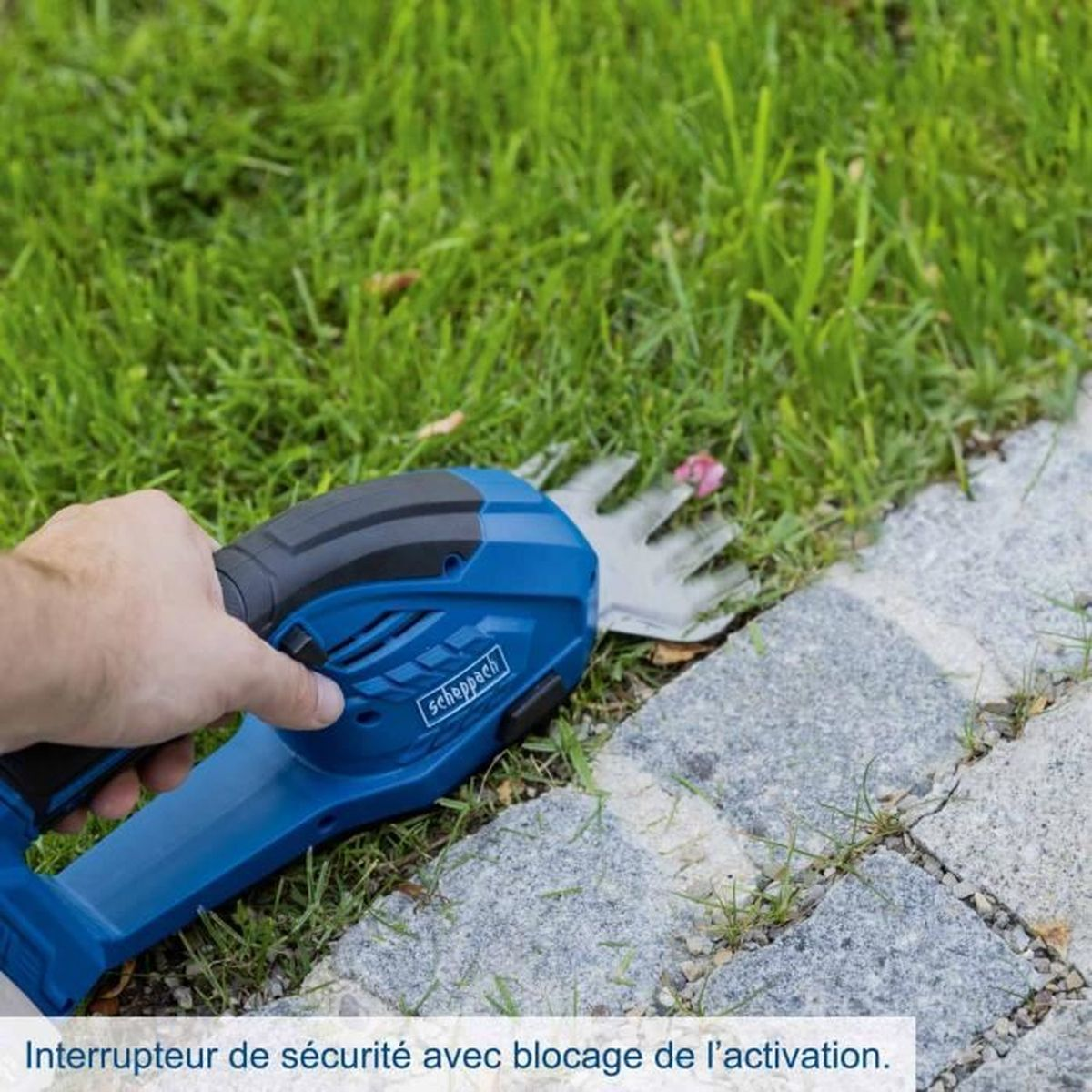 Een hand houdt een blauwe draadloze trimmer vast bij een stenen graskant.