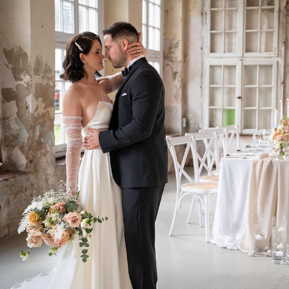 Para młoda w strojach ślubnych tuli się przy oknach; dekoracja stołu z tyłu.