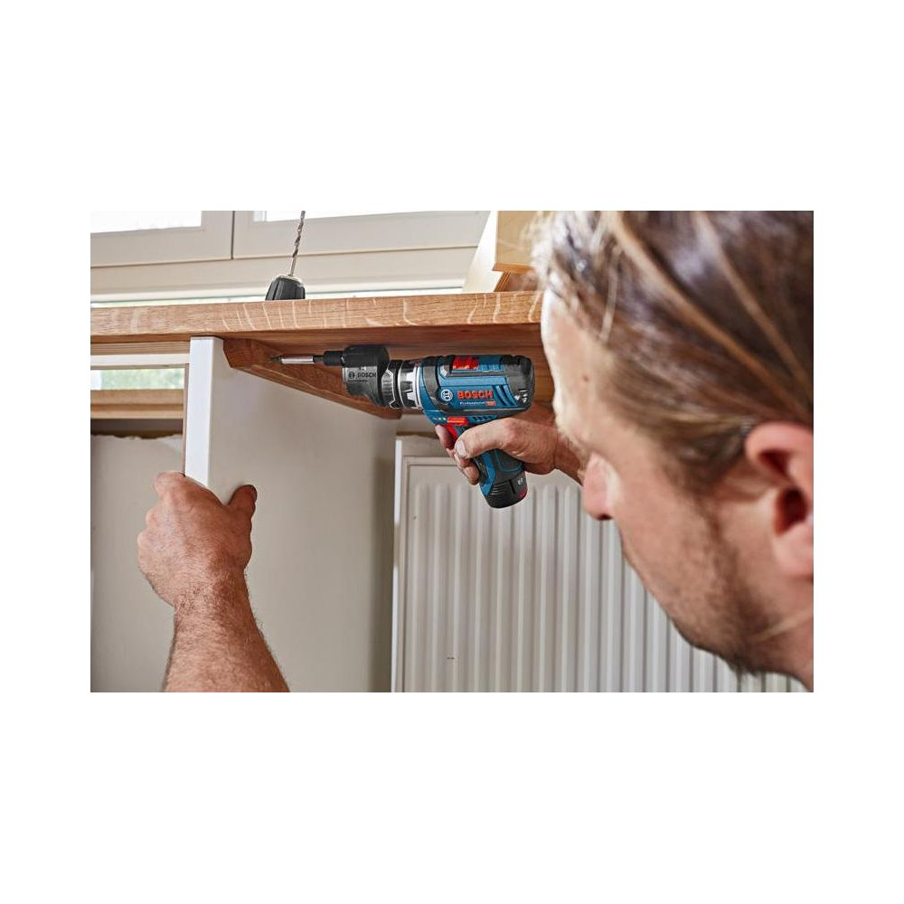 Mężczyzna wierci w drewnie niebieską wiertarką Bosch. Kaloryfer w tle.