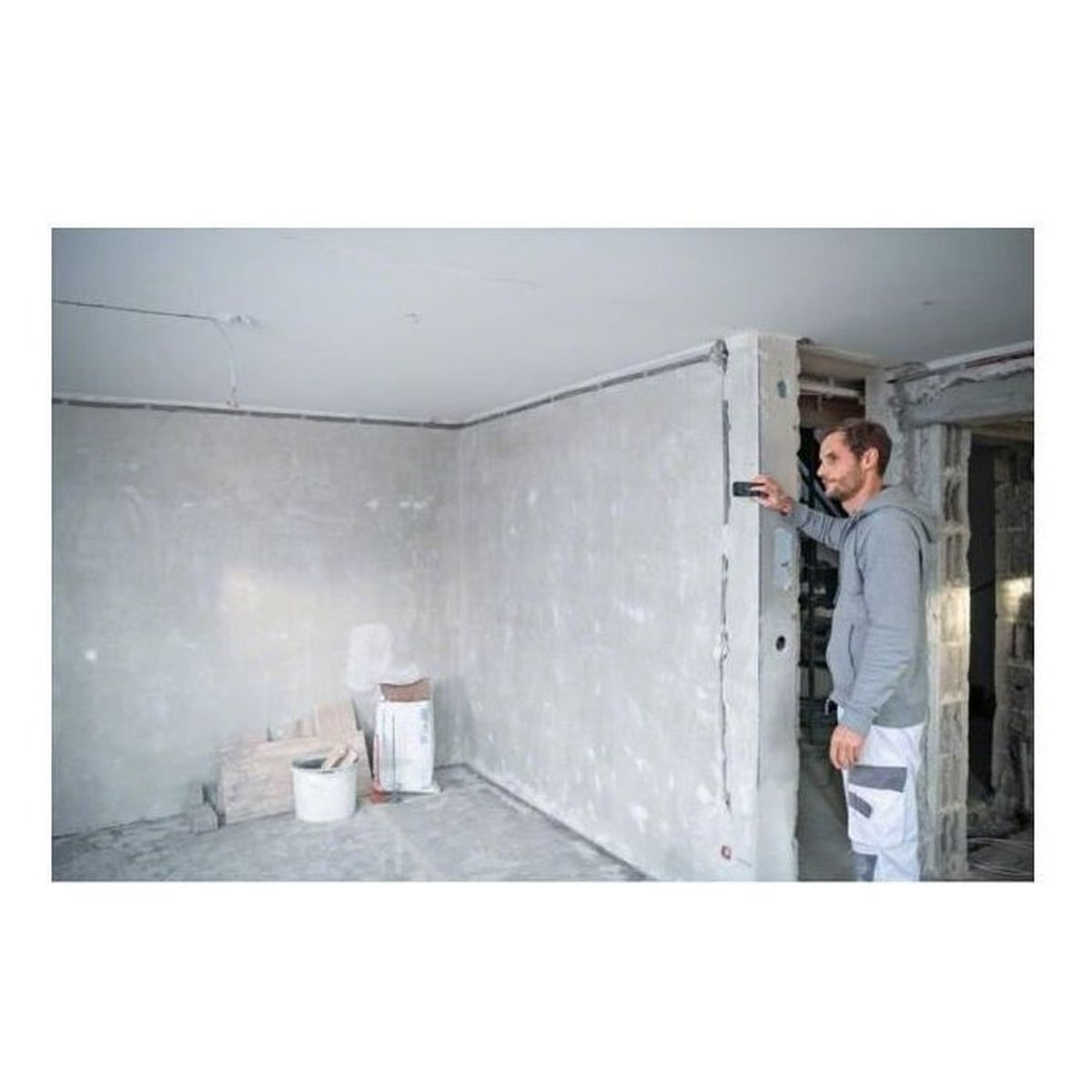 Mann in grauem Kapuzenpullover misst Wand mit Gerät. Baustelle.