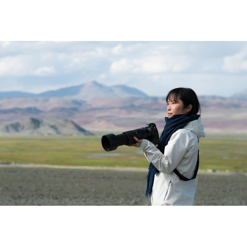 Kobieta z aparatem stoi na rozległym, górzystym terenie.