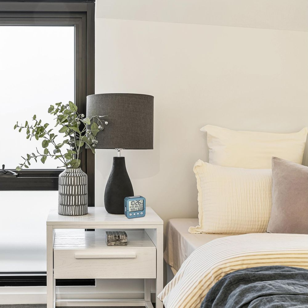 Ein Nachttisch mit Lampe, Vase mit Pflanzen und Digitaluhr neben einem Bett.